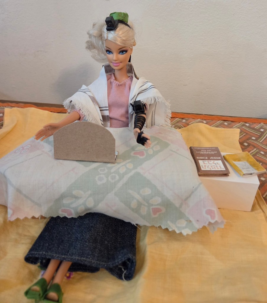 DC Tefillin Barbie (see post for details on her appearance) at a table with a laptop. Talmud volume and siddur are off on a side-table.